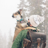 This is a man wearing a dark brown leather backpack in the snow and carrying a wounded dog.